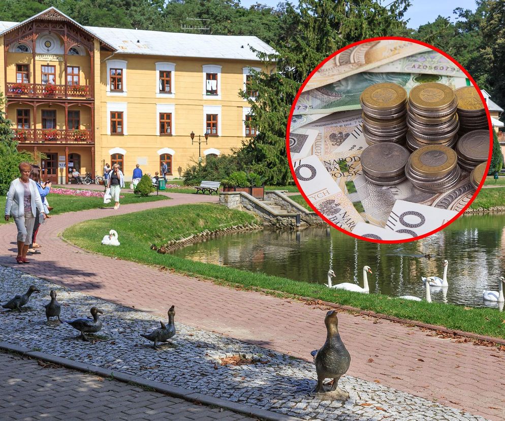 Budynek uzdrowiskowy z drewnianymi zdobieniami i balkonami, otoczony zielenią, z alejką spacerową i stawem, po którym pływają łabędzie. Na pierwszym planie widać spacerujących kuracjuszy i figury gęsi, symbolizujące spokój i odpoczynek. W prawym górnym rogu, w czerwonym okręgu, widoczne są banknoty i monety, obrazujące niższe ceny sanatoryjnych turnusów. Więcej o obniżkach NFZ przeczytasz na Super Biznes.