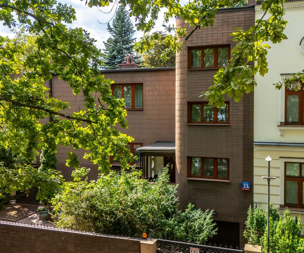 Dom Romualda Gutta przy Kieleckiej 33a w Warszawie, z elewacją pokrytą płytkami, widoczny przez gęste liście drzew, z willą w stylu narodowym po prawej. Więcej o architekturze tego wyjątkowego budynku przeczytasz na Architektura Murator Plus.