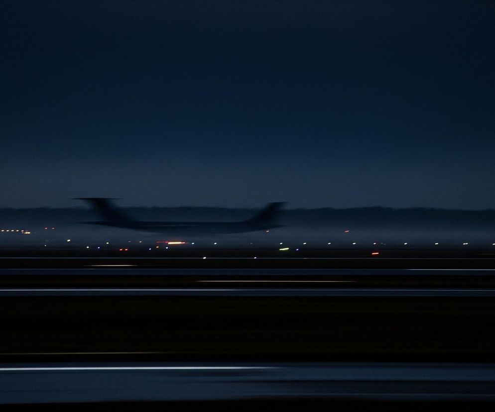 Horyzontalnie rozmyty samolot o ciemnym kadłubie i skrzydłach, widoczny w środkowej części obrazu, znajduje się w ruchu. Tło stanowi ciemne, granatowe niebo, na którym zlewają się gdzieniegdzie jaśniejsze pasma. Poniżej rozmytej sylwetki samolotu rozciąga się pas startowy z licznymi, rozproszonymi światłami w kolorach białym, pomarańczowym i czerwonym, tworzącymi świetlne smugi. Dolna część kadru to ciemny, gładki obszar, prawdopodobnie fragment asfaltowej powierzchni.