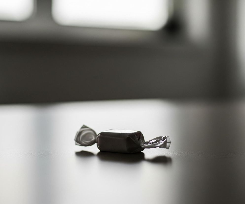 Na ciemnobrązowej, teksturowanej powierzchni leży błyszczący cukierek w opakowaniu. Opakowanie cukierek jest srebrno-szare, z pomarszczonymi końcówkami, a jeden z końców jest lekko rozwinięty. Cukierek rzuca wyraźny cień w prawo, na tle rozmytego, jaśniejszego obszaru w oddali, przypominającego okno. Całość kadru utrzymana jest w stonowanych odcieniach szarości i brązu.