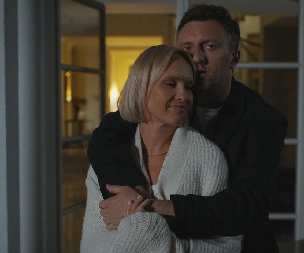 Dorota (Iwona Rejzner) i Bartek (Arkadiusz Smoleński) z serialu M jak miłość przytulają się czule w ogrodzie. Bartek obejmuje Dorotę od tyłu, głaszcząc jej policzek. Para ma na sobie swobodne, wieczorowe stroje. Zdjęcie ilustruje romantyczny prezent Bartka dla Doroty przed wyprowadzką, o czym przeczytasz na portalu Super Seriale.