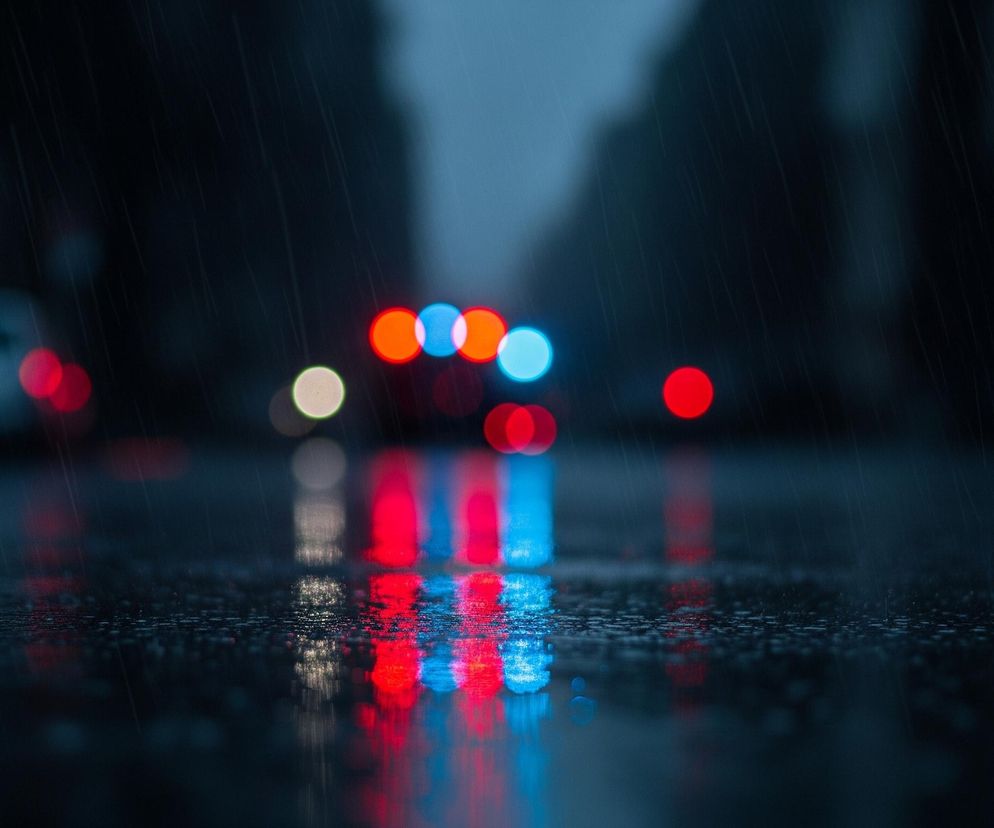 Ciemną, deszczową ulicę, gdzie głównym elementem są rozmyte światła samochodów. Na pierwszym planie widać mokry asfalt z licznymi kroplami deszczu, odbijający czerwone i niebieskie światła z tyłu. W tle, powyżej horyzontu mokrej ulicy, znajdują się okrągłe, rozmazane światła, głównie czerwone i niebieskie, sugerujące jadące pojazdy, a także pojedyncze, blade światło z lewej strony. Całość jest utrzymana w ciemnych, chłodnych barwach, z dominacją granatu i czerni, przełamywanych intensywnymi punktami czerwieni i błękitu.