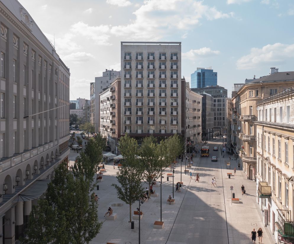 Widok z góry na Plac Pięciu Rogów w Warszawie, ukazujący dużą, wyłożoną płytami przestrzeń publiczną z licznymi przechodniami, drzewami i ławkami. W tle zabytkowe kamienice oraz nowoczesne budynki. Więcej o architekturze miasta na Architektura Murator Plus.