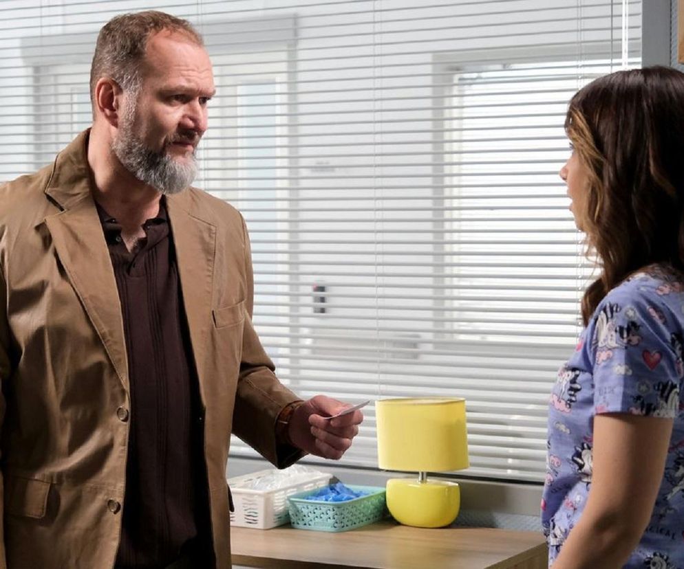 Mirosław Wójcik (Jakub Kornacki) w beżowej marynarce i brązowej koszuli, z siwą brodą, wręcza wizytówkę Anecie Chodakowskiej (Ilona Janyst) w fioletowym fartuchu medycznym. Scena z serialu M jak miłość, o którym możesz przeczytać więcej na Super Seriale.