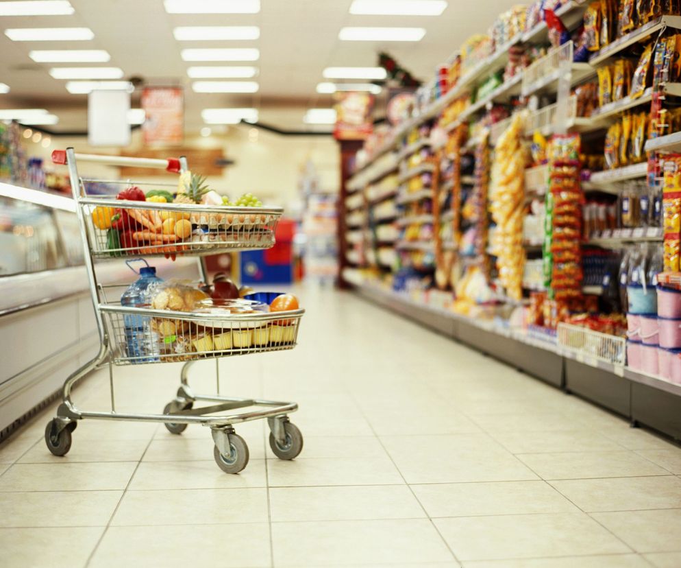 Wózek sklepowy wypełniony różnymi produktami spożywczymi, w tym świeżymi warzywami i owocami, stoi na tle rozmytych półek supermarketu. Ilustracja odzwierciedla kontekst zakupów i bezpieczeństwa żywności, o czym przeczytasz więcej na Poradnik Zdrowie.