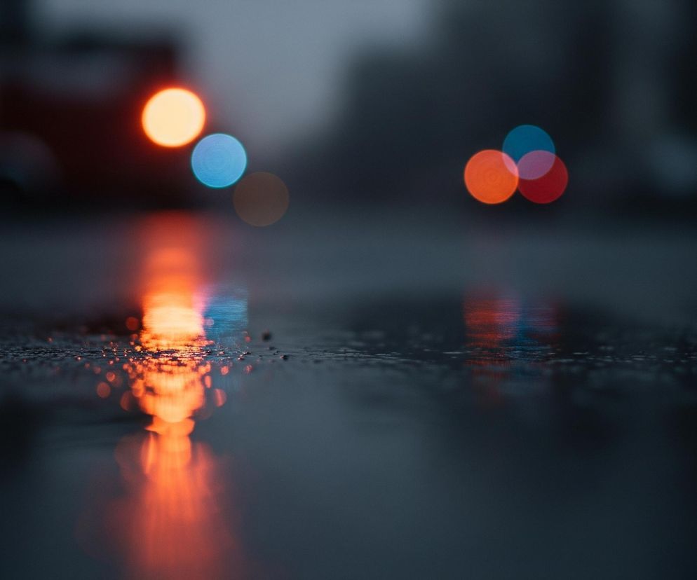 Na pierwszym planie widoczna jest mokra, ciemna nawierzchnia z drobnymi, błyszczącymi kroplami wody, z której odbijają się barwne światła. Głębiej, ponad powierzchnią, unoszą się rozmyte kręgi świateł bokeh w kolorach pomarańczowym, niebieskim i czerwonym, sugerując miejskie otoczenie w deszczową noc. Tło jest zamglone i ciemne, z zarysami budynków lub innych obiektów miejskich.