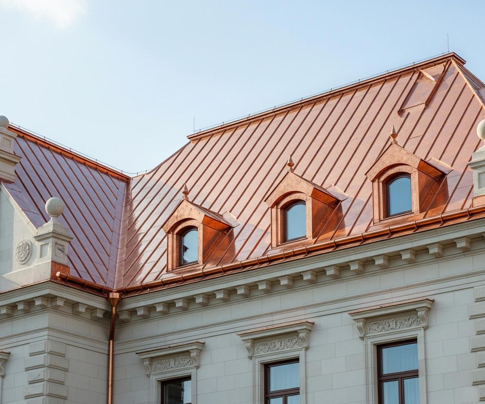 Fragment okazałego budynku z miedzianym dachem i jasną elewacją. Dach, o spadzistej konstrukcji i wyraźnych pionowych liniach, mieni się w odcieniach brązu i pomarańczu, odbijając światło słoneczne. Na nim znajdują się trzy miedziane lukarny z półokrągłymi oknami oraz liczne elementy ozdobne, w tym okrągłe kule umieszczone na szczytach konstrukcji fasadowych. Elewacja jest zrobiona z jasnych, kamiennych bloków, a jej ozdobne detale, takie jak gzymsy, boniowanie narożne i rzeźbione ornamenty nad oknami, podkreślają jej klasyczny styl. W lewej części obrazu widoczne jest rusztowanie, częściowo zasłaniające fragment budynku, co wskazuje na prace konserwatorskie lub budowlane.