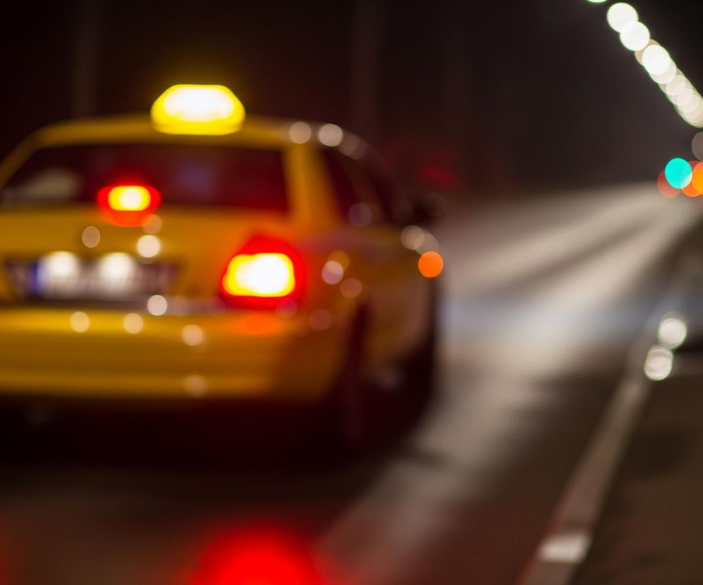 Żółte auto osobowe z pomarańczowym światłem na dachu i włączonymi tylnymi światłami pozycyjnymi oraz światłem stop, jest widoczne od tyłu. Pojazd przemieszcza się po mokrej, ciemnej drodze, z białymi pasami po prawej stronie. Tło jest rozmyte, z widocznymi jasnymi, kulistymi punktami światła o różnych kolorach: białym, zielonym, pomarańczowym i czerwonym, sugerującymi nocną scenerię miejską.