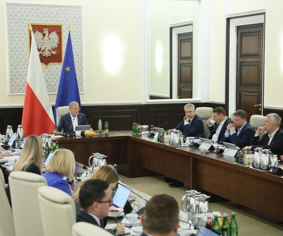 Donald Tusk przewodniczący posiedzeniu rządu w otoczeniu polityków, z biało-czerwoną flagą i flagą Unii Europejskiej w tle. Wszyscy ubrani w formalne stroje, z uwagą śledzący obrady. O kluczowej ustawie o asystencji osobistej przeczytasz na Super Biznes.