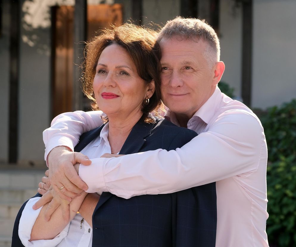 Małgorzata Pieńkowska (Marysia) i Robert Moskwa (Artur) z serialu M jak miłość na zdjęciu, gdzie Artur obejmuje Marysię od tyłu. Uśmiechnięci Rogowscy stoją na tle zieleni i budynku. Więcej o ich losach w M jak miłość przeczytasz na Super Seriale.