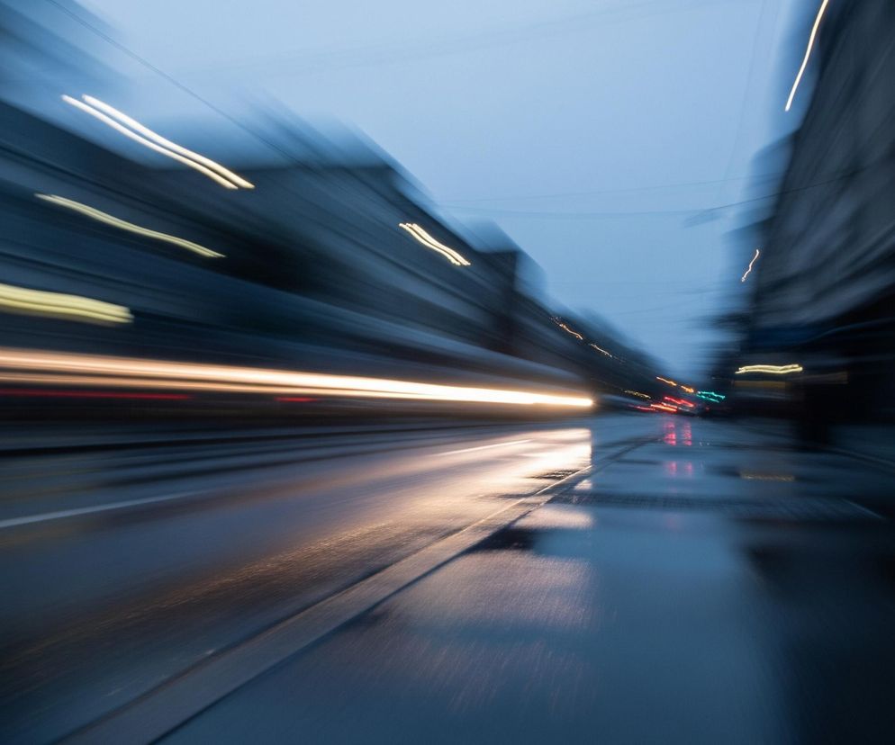 Rozmyty obraz przedstawia nocny widok ulicy, z widocznymi smugami światła wskazującymi na ruch. Na lewej stronie widać długą, jasną smugę światła biegnącą poziomo, sugerującą przejeżdżający pojazd, której blask odbija się na mokrej nawierzchni drogi. Po obu stronach drogi rozciągają się ciemne, niewyraźne kształty budynków, z kilkoma pionowymi smugami żółtego światła w górnej części kadru. Niebo jest ciemnoniebieskie, a mokra jezdnia odbija otaczające światła.
