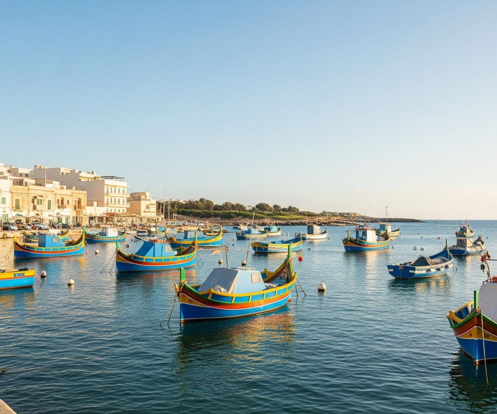 W płytkiej, spokojnej zatoce, pełnej jaskrawych, wielokolorowych łodzi, dominują odcienie błękitu i żółci. Na horyzoncie, po lewej stronie, rozciąga się linia białych i beżowych budynków, oświetlonych ciepłym światłem. Woda odbija błękit nieba, tworząc faliste refleksy wokół zakotwiczonych jednostek. Kamienny brzeg z pachołkami i linami widoczny jest w lewej dolnej części obrazu.