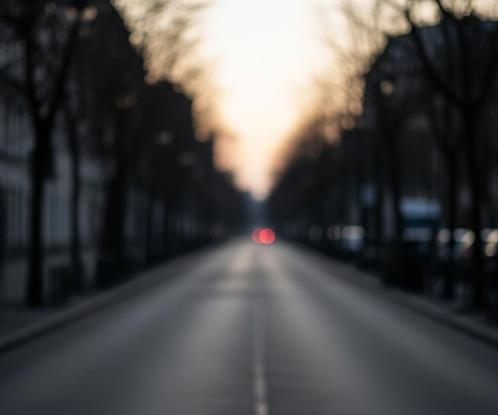 Długa, prosta ulica, po obu stronach której rosną drzewa oraz stoją budynki, rozciąga się w kierunku odległego, jasnego punktu. Droga jest szara z niewyraźną białą linią centralną, a na jej końcu widoczne są dwa rozmyte, czerwone światła. Całość jest nieostra, tworząc efekt bokeh, z ciemnymi, niewyraźnymi sylwetkami drzew i budynków wzdłuż krawędzi kadru.