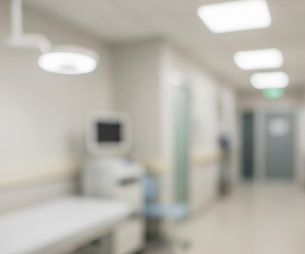 Rozmyty obraz przedstawia jasny korytarz lub pomieszczenie medyczne, z dominującymi odcieniami bieli i beżu. Po lewej stronie widać leżankę medyczną z białym obiciem oraz część nieostrego sprzętu medycznego z monitorem na górze. Sufit jest oświetlony prostokątnymi i okrągłymi lampami, a dalej w głąb korytarza widoczne są drzwi oraz fragmenty ścian w jaśniejszych barwach.