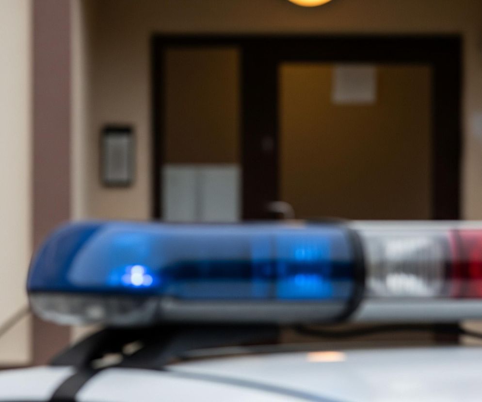 Na pierwszym planie widoczny jest górny element pojazdu, prawdopodobnie samochodu, z charakterystycznymi światłami sygnalizacyjnymi. Dominują tu światła koloru niebieskiego po lewej stronie i czerwonego po prawej, oba intensywnie świecące i nieco rozmyte, co sugeruje ruch lub fotografowanie z małą głębią ostrości. Tło jest całkowicie rozmyte, przedstawiając fasadę budynku w odcieniach beżu i brązu, z ciemnymi prostokątnymi kształtami drzwi lub okien.