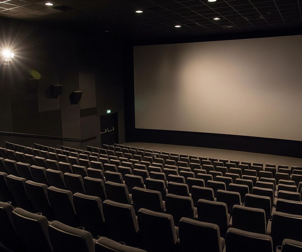 Pusta sala kinowa z rzędami szarych foteli rozciągających się od lewej dolnej części obrazu ku górze i prawej. Na wprost widoczny jest duży, prostokątny ekran projekcyjny o jasnoszarym odcieniu, zajmujący większą część ściany. Z lewej strony, w górnej części obrazu, intensywne światło z projektora rzuca długie smugi i flary, oświetlając lekko część ekranu. Cała sala, łącznie ze ścianami i sufitem, utrzymana jest w ciemnych barwach, głównie czerni i szarości, co kontrastuje z jasnym ekranem i punktowym światłem projektora.