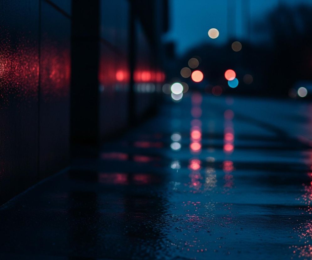 Ściana w ciemnym odcieniu granatu z widocznymi, drobnymi kroplami wody, odbijającymi intensywne czerwone światło, zajmuje lewą stronę kadru. Niżej znajduje się mokra nawierzchnia chodnika lub ulicy, również pokryta drobnymi kroplami i odbijająca rozmyte światła w różnych kolorach. Dalej, w tle, widoczna jest rozmyta scena miejska z okrągłymi światłami w odcieniach czerwieni, bieli, pomarańczu i niebieskiego, sugerująca nocny ruch uliczny lub sygnalizację. Całość obrazu utrzymana jest w chłodnej tonacji granatu i błękitu, z dominującymi akcentami czerwonego światła.