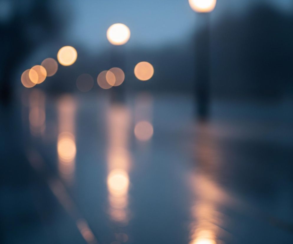 Rozmyte ujęcie ulicy nocą, gdzie widoczne są jaskrawe, okrągłe plamy światła latarni ulicznych, odbijające się na mokrej nawierzchni. Kolorystyka obrazu jest zdominowana przez ciemne odcienie niebieskiego i szarości, kontrastujące z ciepłymi, pomarańczowo-żółtymi punktami świetlnymi. Latarnie ułożone są w perspektywie, prowadzącej wzrok w głąb obrazu, a ich odbicia tworzą smugi światła na mokrej drodze, sugerując deszcz lub świeżo po nim.