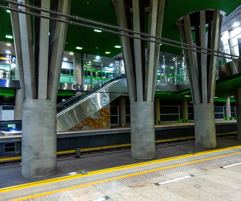 Wnętrze stacji metra Stadion Narodowy w Warszawie z peronem linii M3, z masywnymi kolumnami wspierającymi zielony sufit, srebrnymi ruchomymi schodami oraz sklepami na antresoli. Więcej o planach i budowie 3. linii metra przeczytasz na Architektura Murator Plus.