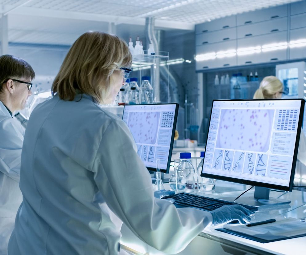 Laboratorium badawcze, gdzie grupa naukowców w białych kitlach analizuje dane na monitorach komputerowych. Na ekranach widoczne są grafiki DNA i obrazy komórek. W tle widać kolby i sprzęt laboratoryjny. Na temat przełomowych badań dowiesz się więcej na Poradnik Zdrowie.