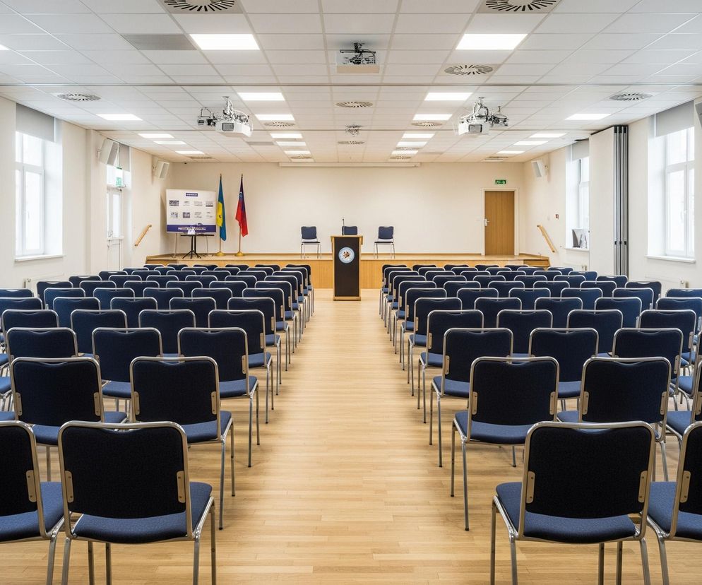W przestronnej sali konferencyjnej, oświetlonej równomiernie lampami panelowymi umieszczonymi w podwieszanym suficie, rzędy krzeseł z ciemnoniebieskimi tapicerowanymi siedzeniami i oparciami oraz srebrnymi, metalowymi ramami, są ustawione symetrycznie na jasnej, drewnianej podłodze, tworząc dwie główne sekcje przed podwyższoną sceną. Na scenie znajduje się ciemnobrązowa mównica z godłem, dwie puste krzesła z niebieskimi siedzeniami oraz dwa słupy z flagami: jedna o barwach żółto-niebiesko-czerwonych, a druga niebiesko-żółto-niebieskich. Za mównicą stoi stół z jasnym obrusem, a obok niego, po lewej stronie sceny, widoczna jest biała tablica informacyjna z tekstami i grafikami. Na ścianach po obu stronach sali znajdują się duże okna z białymi ramami i roletami, a także głośniki i otwory wentylacyjne w suficie.