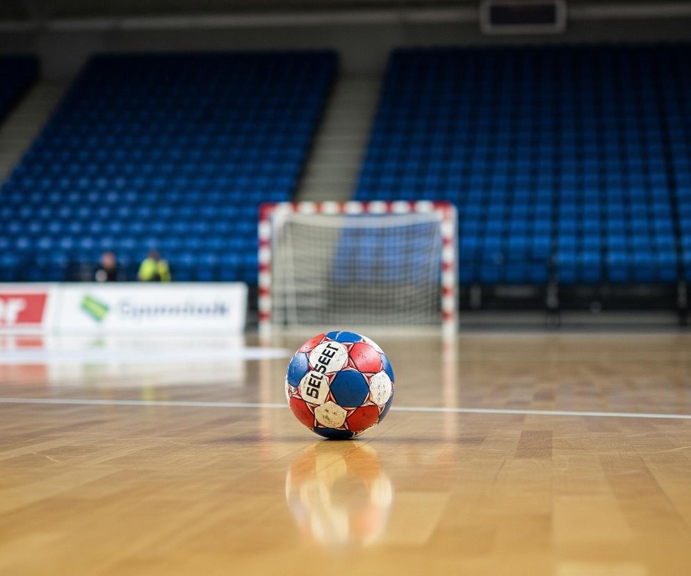 Piłka do piłki ręcznej leży na jasnobrązowej, lakierowanej podłodze boiska sportowego. Jest ona czerwono-niebiesko-biała, z napisem SELECT widocznym na białych panelach. Tło stanowi rozmyta trybuna z rzędami niebieskich krzesełek, a w oddali widać bramkę sportową z czerwono-białą obramówką i siatką.