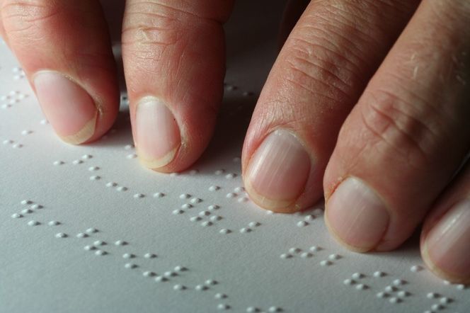 Palce delikatnie przesuwają się po białej kartce z wypukłymi literami Braille'a. Obraz symbolizuje oszustwo niewidomego z Włoch, który przez 50 lat udawał niewidomego, wyłudzając świadczenia. Więcej o tej sprawie przeczytasz na portalu Super Biznes.