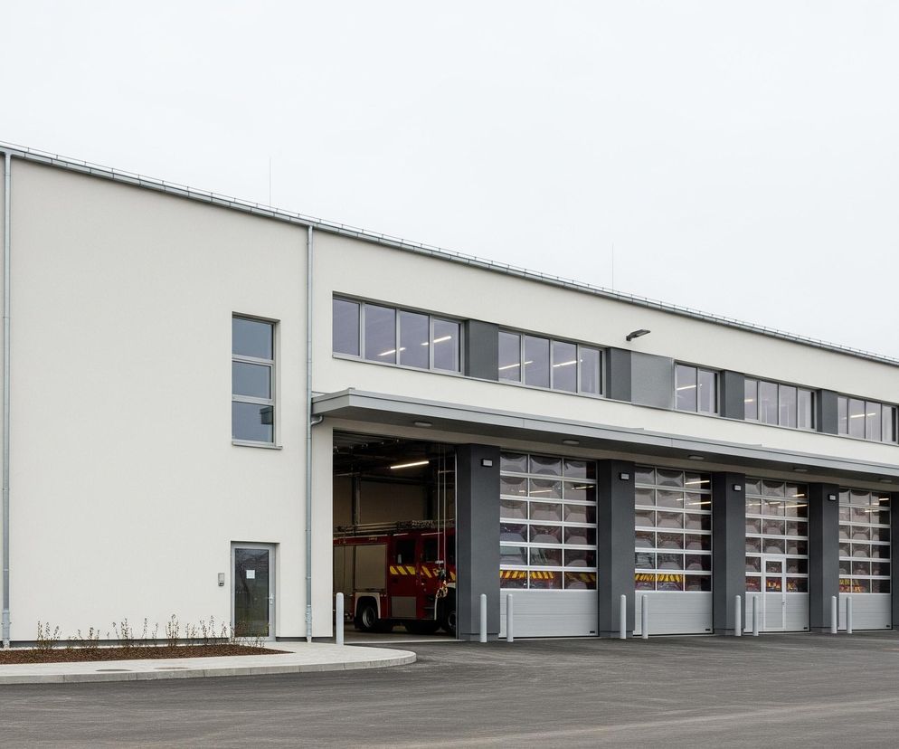 Nowoczesny budynek w kształcie prostopadłościanu, otynkowany na biało i ciemnoszaro, dominuje nad szerokim, szarym placem. Jego fasada składa się z czterech dużych, przeszklonych bram garażowych z dolnymi panelami w kolorze jasnoszarym, z których jedna jest częściowo otwarta, ukazując fragment czerwonego wozu strażackiego z żółtymi elementami. Powyżej bram znajdują się rzędy poziomych okien z ciemnymi szybami, a cała konstrukcja zwieńczona jest płaskim dachem z rynną. Po lewej stronie fasady umieszczone są mniejsze okna i szklane drzwi wejściowe, obok których znajduje się młode drzewo z podporami, a w oddali widoczna jest siatka ogrodzeniowa i nagie drzewa.