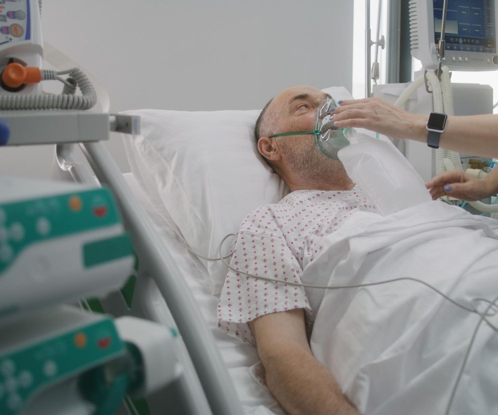 Pacjent z maską tlenową leżący na szpitalnym łóżku, pielęgniarka poprawia mu maskę. Obok widoczne urządzenia medyczne, mierzące funkcje życiowe. Dowiedz się więcej o falach chorób w Polsce i profilaktyce infekcji na Poradnik Zdrowie.