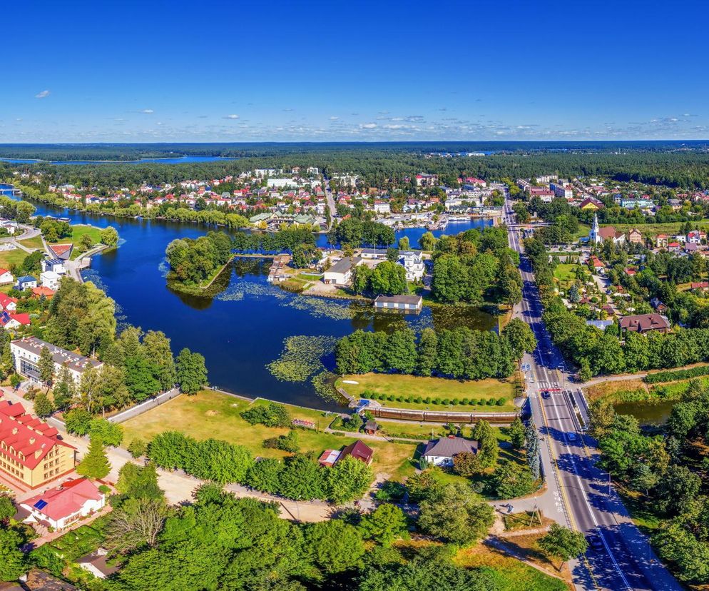 Panoramiczny widok z lotu ptaka na Augustów, turystyczną perłę województwa podlaskiego. Widać malownicze jeziora z wodnymi roślinami, kanały, zielone tereny, zabudowania mieszkalne i ulice z samochodami. Więcej o atrakcjach turystycznych przeczytasz na naszym portalu.