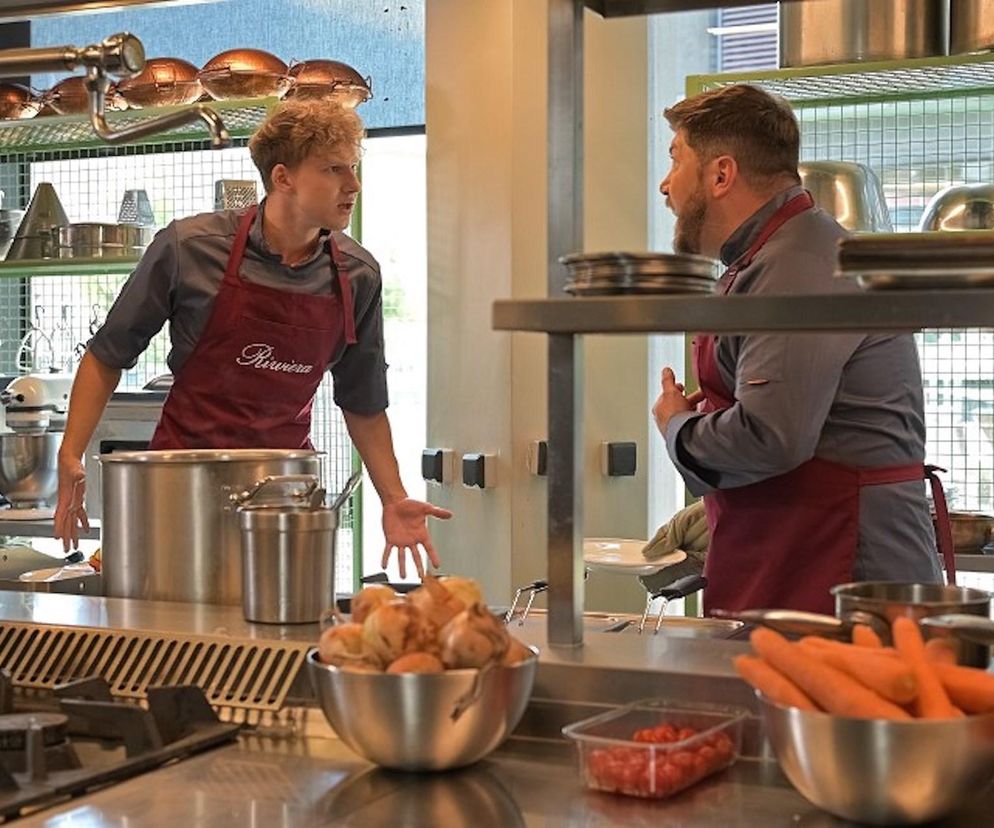 Aktor Oskar Stoczyński jako Tomek w kuchni restauracji, w czerwonym fartuchu z napisem Riviera, rozmawia z menadżerem, również w kucharza stroju, w dynamicznej scenie z serialu Pierwsza miłość, gdzie Tomek jest oskarżony o kradzież. Więcej o dramacie Tomka znajdziesz na Super Seriale.