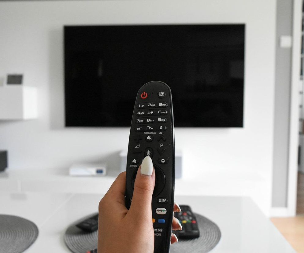 Dłoń kobiety w białym manicure trzymająca czarny pilot do telewizora ze srebrnymi przyciskami, z telewizorem z czarnym ekranem w tle, ilustrująca niedosłuch i problem głośniejszego oglądania telewizji. Dowiedz się więcej o zdrowiu mózgu na Poradnik Zdrowie.