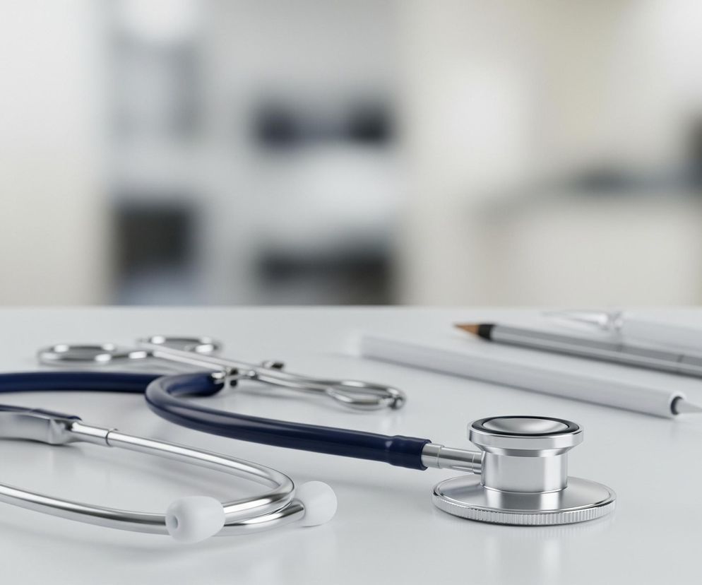 Na białym blacie stołu leży granatowy stetoskop z metalowymi elementami i białymi oliwkami. Obok niego, częściowo ukryte, znajdują się srebrne nożyczki chirurgiczne. Dalej w tle widać trzy długopisy: dwa białe, z czego jeden ma niebieską skuwkę, oraz jeden czarny. Tło jest rozmyte, o jasnych, beżowo-szarych barwach, sugerujących wnętrze pomieszczenia.