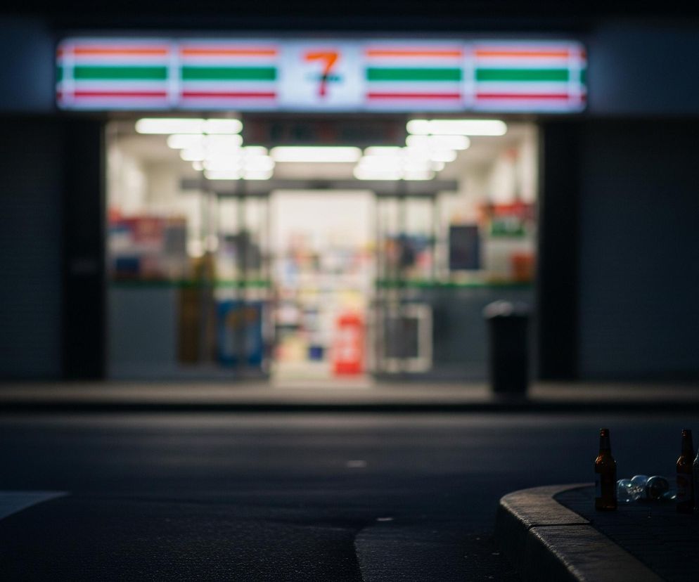 Na zdjęciu dominuje ciemna, nocna sceneria. W oddali, pośrodku kadru, widoczny jest jasno oświetlony sklep. Jego fasada jest ciemna, ale nad wejściem znajduje się podświetlany szyld z napisem 7 ELEVEN w charakterystycznych czerwono-pomarańczowo-zielonych pasach. Wnętrze sklepu jest bardzo jasne, oświetlone białym światłem jarzeniówek, co tworzy kontrast z ciemnym otoczeniem.

Na pierwszym planie, po prawej stronie, leży kilka pustych butelek po napojach, w tym dwie brązowe butelki oraz jedna przezroczysta, szklana butelka. Butelki leżą na ciemnym chodniku, tuż przy krawężniku. Fragment śmietnika z białą, foliową torbą wystającą z góry jest widoczny z prawej strony. Asfalt ulicy na pierwszym planie jest ciemny i gładki, z delikatnymi refleksami światła odbijającymi się od powierzchni.