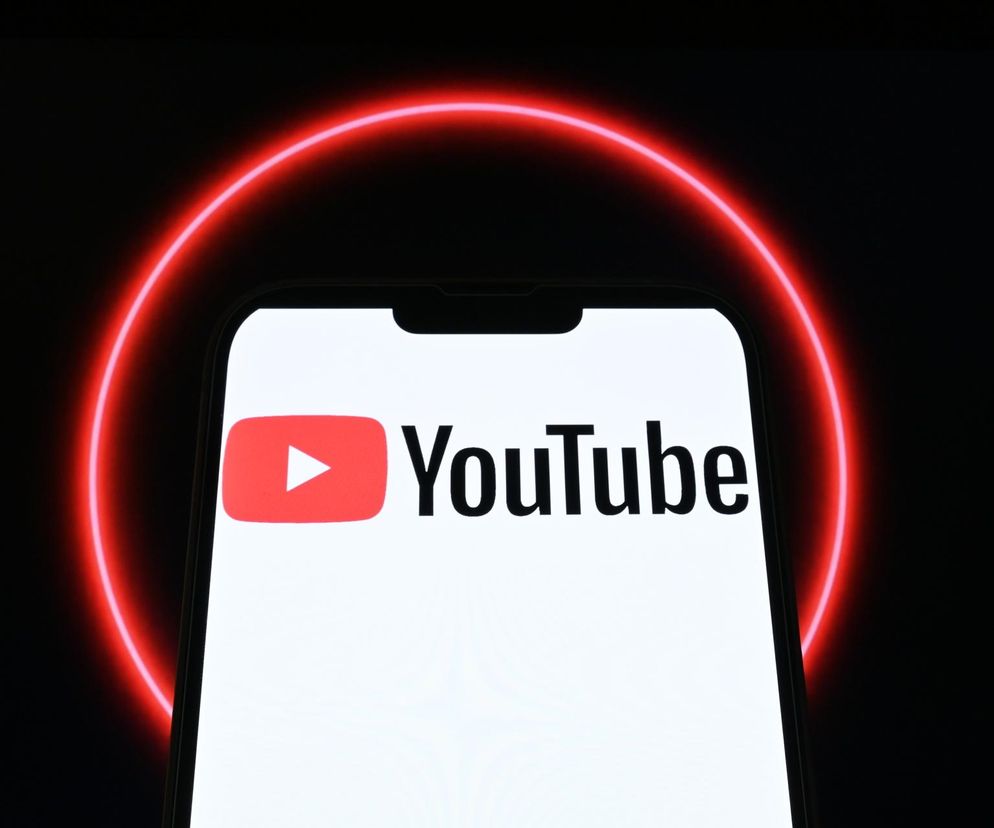 Smartfon z otwartą aplikacją YouTube, logo platformy wyraźnie widoczne na białym ekranie, otoczone neonową czerwoną poświatą symbolizującą dynamiczny rozwój i globalny zasięg. O nowych funkcjach przeczytasz na Super Biznes.