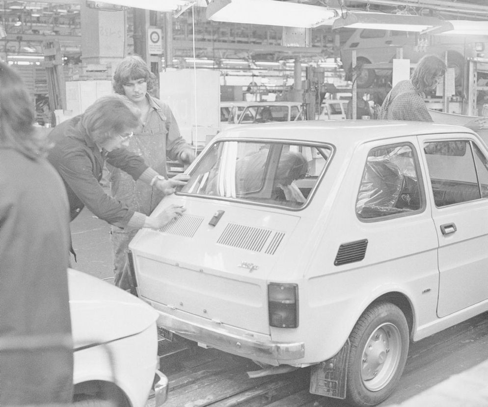 Pracownicy montujący tylną szybę w białym Fiacie 126p na linii produkcyjnej fabryki FSM. Scena z epoki PRL, ukazująca proces produkcji kultowego malucha, który zmotoryzował Polskę. Więcej o historii tego pojazdu na portalu Super Biznes.