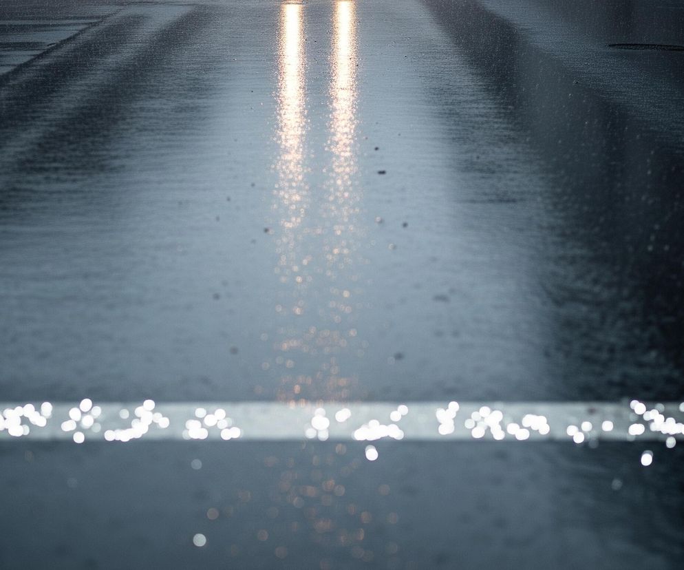 Mokra, ciemnoszara nawierzchnia drogi z widocznymi odbiciami świateł zajmuje większą część kadru. Dwie pionowe, jasne smugi światła o pomarańczowo-żółtym odcieniu odbijają się na mokrej powierzchni w górnej części obrazu, sugerując światła pojazdu. W dolnej części zdjęcia biegnie pozioma, biała linia, która jest rozmyta i składa się z wielu jasnych, okrągłych punktów świetlnych, imitujących efekt bokeh. Pozostała część drogi jest jednolita, ciemna i błyszcząca, z delikatnymi, nieregularnymi refleksami.