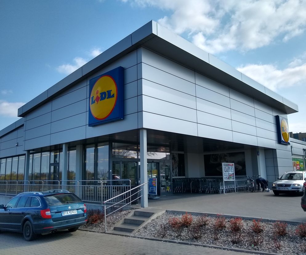 Supermarket Lidl z parkingiem i dwoma samochodami, w tym granatową Skodą. Przed wejściem widoczne rzędy wózków sklepowych. Obraz odzwierciedla temat niedziel handlowych, o których można przeczytać więcej na Super Biznes.