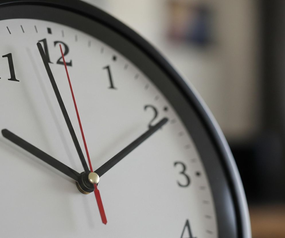 Zbliżenie przedstawia tarczę zegara ściennego z czarną ramką i białym tłem. Czarne wskazówki godzinowa i minutowa wskazują pozycję bliską godziny dziesiątej, zaś cienka, czerwona wskazówka sekundowa znajduje się tuż przed godziną dwunastą. Na tarczy widoczne są czarne cyfry od 9 do 2, oraz krótkie, czarne kreski oznaczające minuty i godziny. W centralnej części tarczy, gdzie stykają się wskazówki, znajduje się mały, złoty element. Tło za zegarem jest nieostre, w odcieniach beżu, bieli i brązu, sugerując wnętrze pomieszczenia.