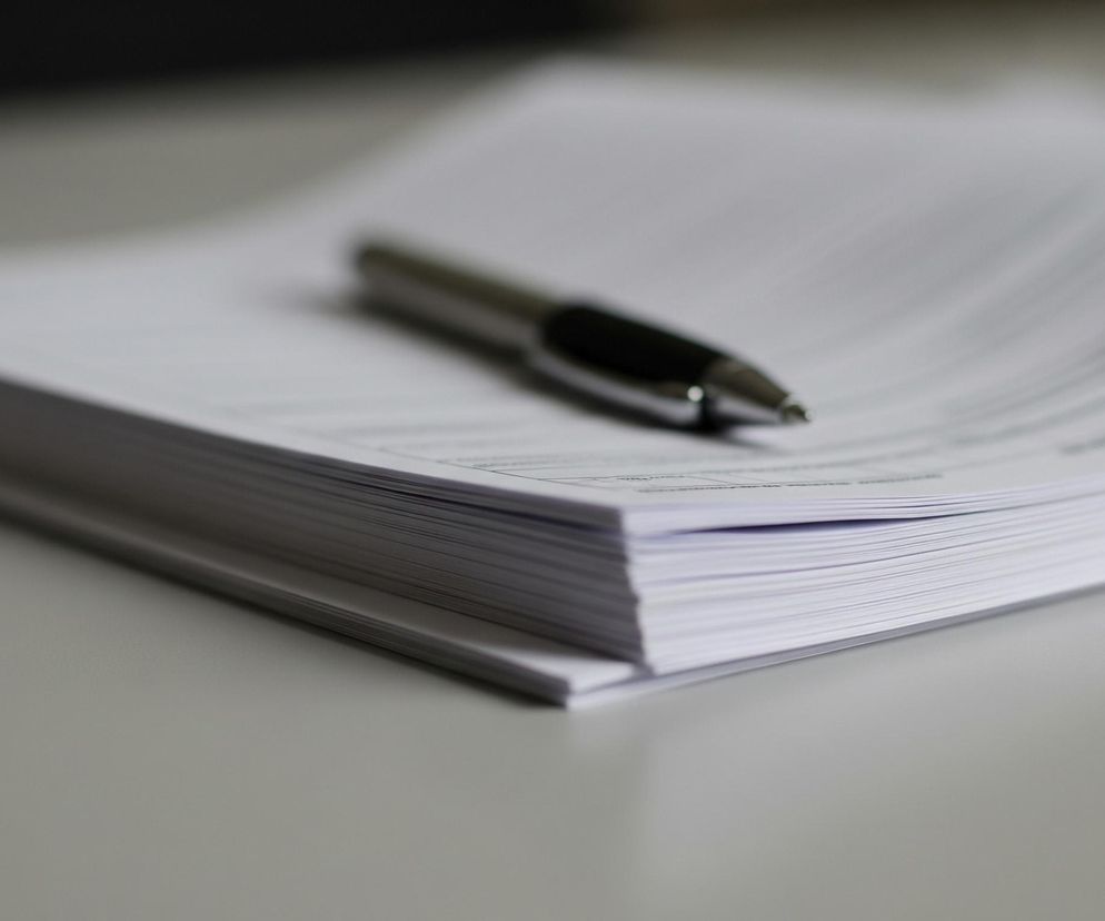 Na blacie stołu widać stos białych kartek papieru z delikatnie zaokrąglonymi rogami, ułożonych ukośnie, z lewego dolnego rogu do prawego górnego. Na wierzchu sterty leży długopis z czarnym korpusem i srebrnymi elementami, skierowany w prawą stronę. Tło obrazu jest nieostre, widoczne są jedynie rozmyte ciemniejsze obszary u góry, kontrastujące z jasnym blatem.