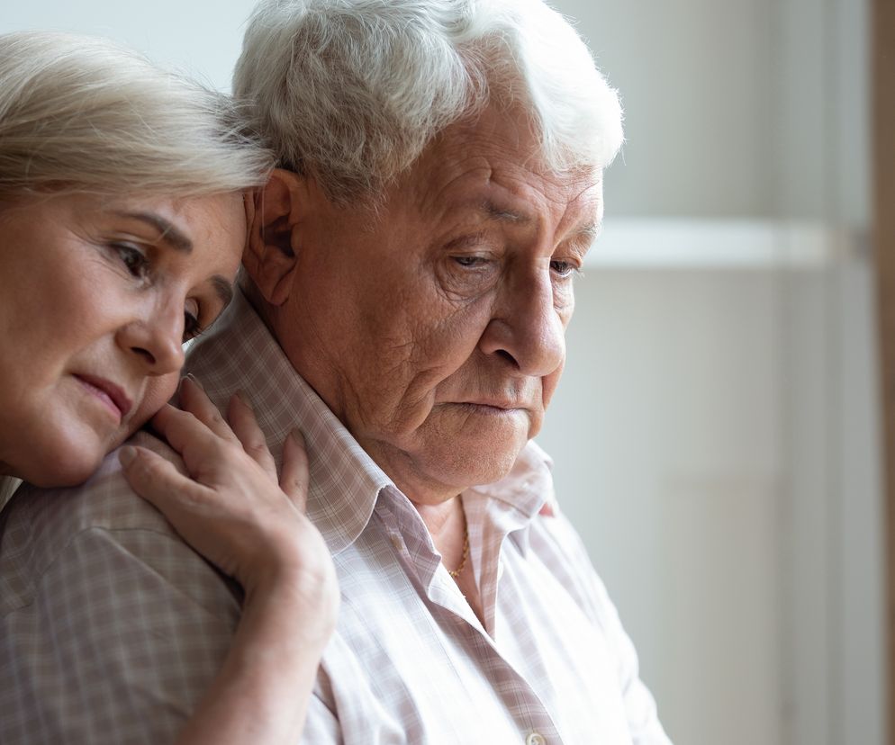 Kobieta o jasnych włosach, wyrażająca współczucie, opiera głowę na ramieniu smutnego, starszego mężczyzny z siwymi włosami, symbolizując wsparcie w obliczu demencji. Artykuł na Poradnik Zdrowie omawia związek depresji i lęku z ryzykiem demencji.