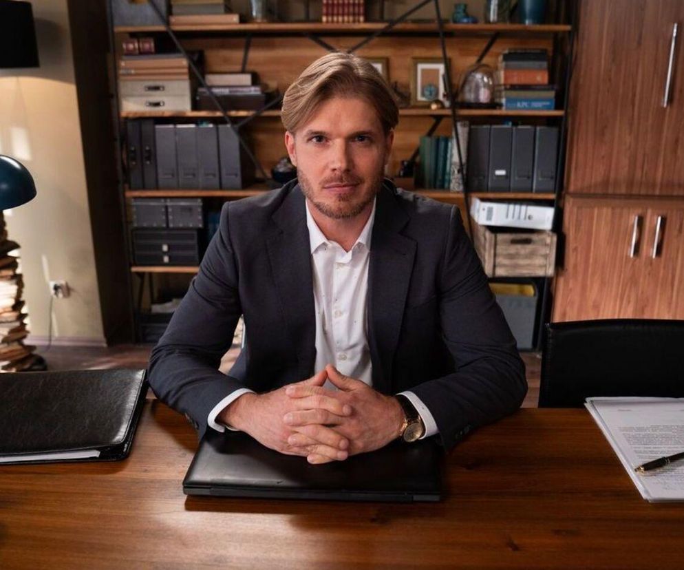 Maks Beger (Tomasz Ciachorowski) w garniturze siedzi za biurkiem, opierając splecione dłonie na laptopie. Na drewnianym biurku widoczne są dokumenty, czarny notes oraz lampa. W tle regał z książkami i segregatorami. Wszystko to można zobaczyć na portalu Super Seriale.