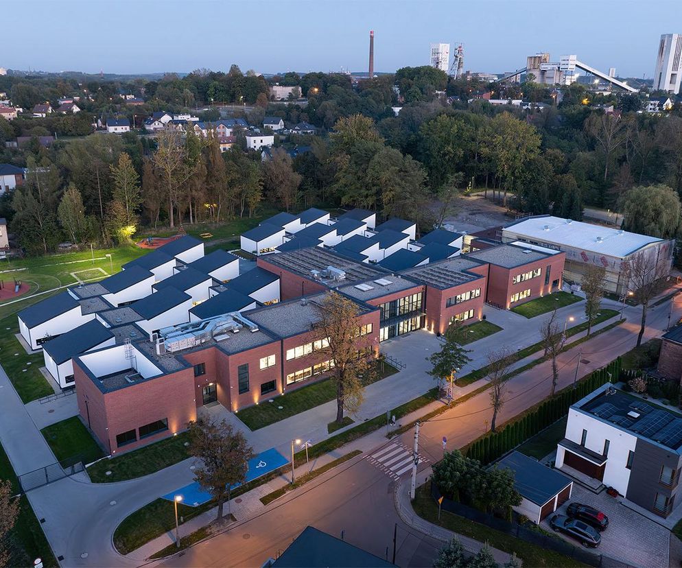 Modułowa Szkoła Podstawowa w Rudzie Śląskiej autorstwa Hornik Chmura Architektura i TKHolding Truszczyński Kobierzewski, widoczna z lotu ptaka o zmierzchu, z ceglanymi elewacjami i białymi dachami na tle zieleni. W tle zabudowania mieszkalne i przemysłowe. Realizację docenioną na łamach Architektura Murator Plus wyróżnia adaptacja do szkód górniczych.