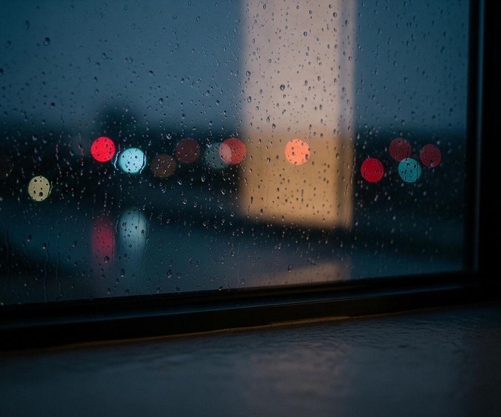 Główne ujęcie to mokra od deszczu szyba z wyraźnymi kroplami wody, za którą widać rozmyte, kolorowe światła. Na pierwszym planie, w dolnej części obrazu, znajduje się parapet lub płaska powierzchnia w ciemnym odcieniu. Dalej widoczna jest czarna rama okienna, a za szybą rozciąga się nieostry widok na miasto nocą lub o zmierzchu, z dominującymi odcieniami niebiesko-szarymi. Spośród rozmytych świateł wyróżniają się okrągłe plamy koloru czerwonego, turkusowego, żółtego i pomarańczowego.