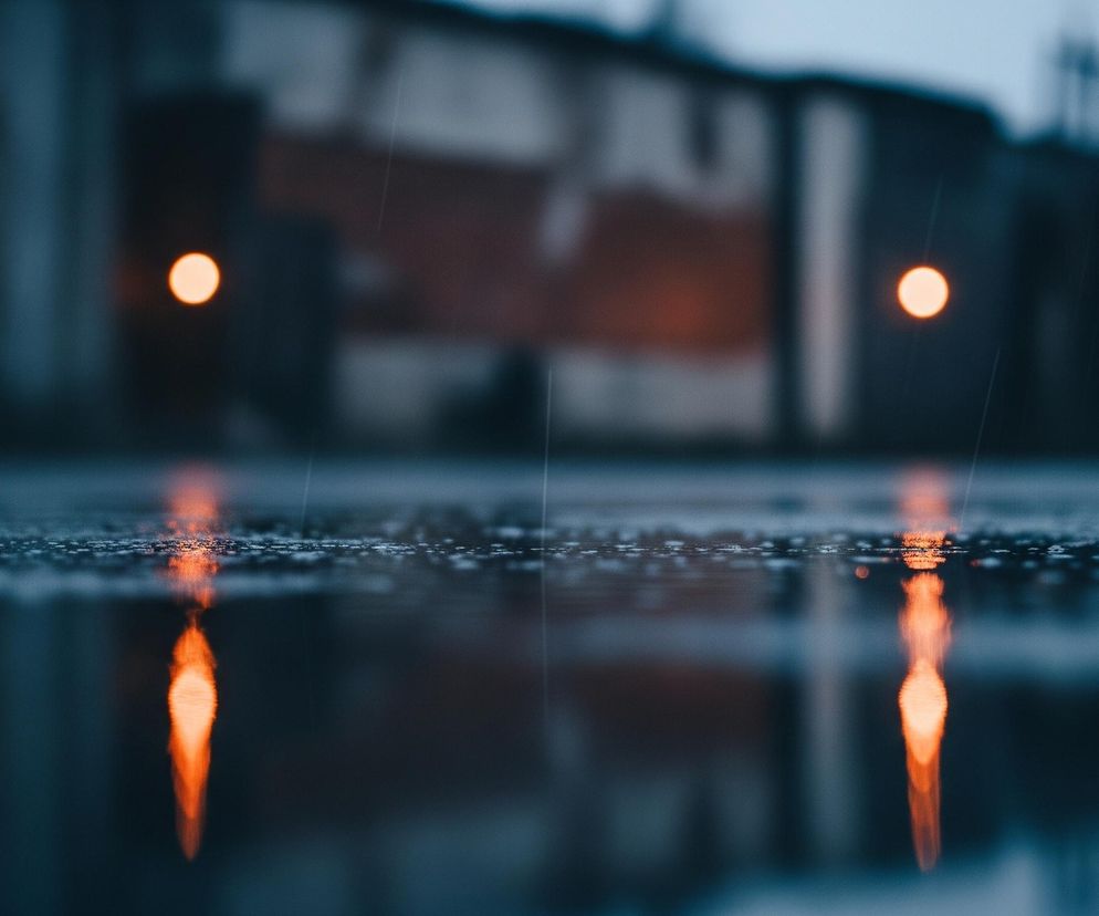 Powierzchnia asfaltu pokryta kroplami deszczu z licznymi odbiciami światła tworzy pierwszy plan, rozciągający się od dołu do około połowy wysokości kadru. Na mokrej nawierzchni widoczne są pomarańczowe, rozmyte odbicia dwóch okrągłych świateł, które znajdują się w tle. Tło jest nieostre i przedstawia zarysy budynków w szarogranatowych odcieniach, z dwoma wyraźnymi, jasnopomarańczowymi punktami świetlnymi, symetrycznie rozmieszczonymi po lewej i prawej stronie. Delikatne, białe smugi deszczu przecinają całą kompozycję.