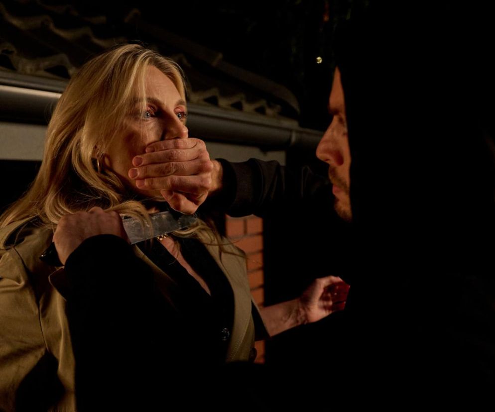 Monika Cieślikowa (Sylwia Gliwa) z blond włosami, ubrana w beżową marynarkę, z nożem przy szyi i zakrytymi ustami przez rękę Bruna (Dawida Czupryńskiego), który stoi obok niej w czarnej bluzie z kapturem. Scena z serialu Na Wspólnej w ciemnym otoczeniu. Więcej o ich relacji przeczytasz na Super Seriale.