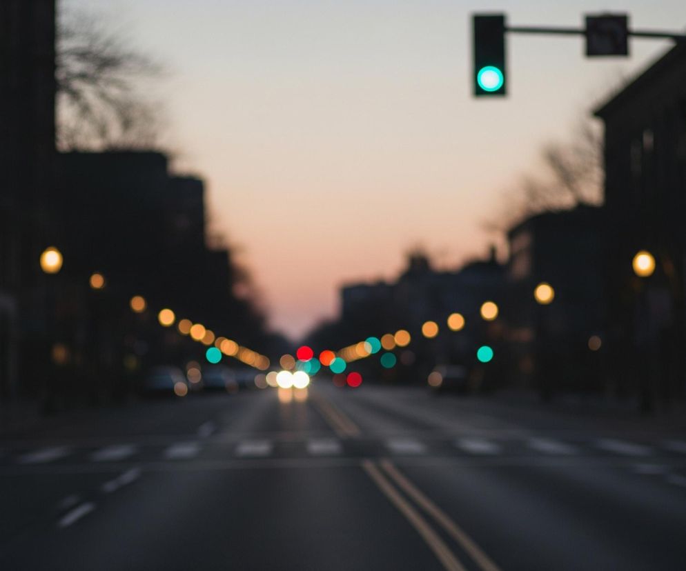 W centrum kadru biegnie asfaltowa droga z dwoma żółtymi pasami wyznaczającymi jej środek, rozciągająca się w kierunku horyzontu. Po obu stronach drogi widać ciemne sylwetki budynków i drzew, które wtapiają się w pastelowe, rozmyte niebo o odcieniach pomarańczy i błękitu. W oddali, wzdłuż drogi, widoczne są liczne, rozmyte światła: pomarańczowe latarnie uliczne, czerwone i zielone światła sygnalizacji oraz jasne reflektory nadjeżdżających samochodów, tworzące efekt bokeh. Na pierwszym planie po prawej stronie znajduje się niewyraźne pomarańczowe światło sygnalizacji, a w górnej części kadru po lewej stronie dominuje zielone światło sygnalizacji.