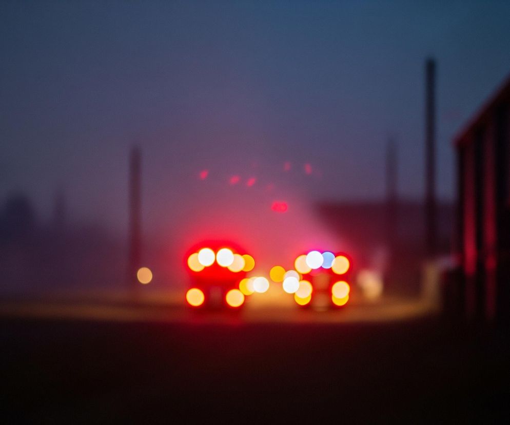 Dwa rozmyte, intensywnie czerwone światła, przypominające tylne światła pojazdu, dominują centralną część zdjęcia, otoczone mniejszymi, jaśniejszymi punktami świetlnymi w odcieniach pomarańczu i bieli. Cała scena jest niewyraźna, z dominującymi barwami ciemnego granatu i fioletu w tle, które płynnie przechodzą w czarny dół obrazu. Po prawej stronie widać zarys ciemnej struktury budowlanej, a w oddali po lewej sylwetki wysokich, pionowych obiektów. Światła tworzą bokeh, a ich otoczenie jest spowite lekko mglistym, niebiesko-fioletowym blaskiem, rozjaśniającym przestrzeń wokół nich.