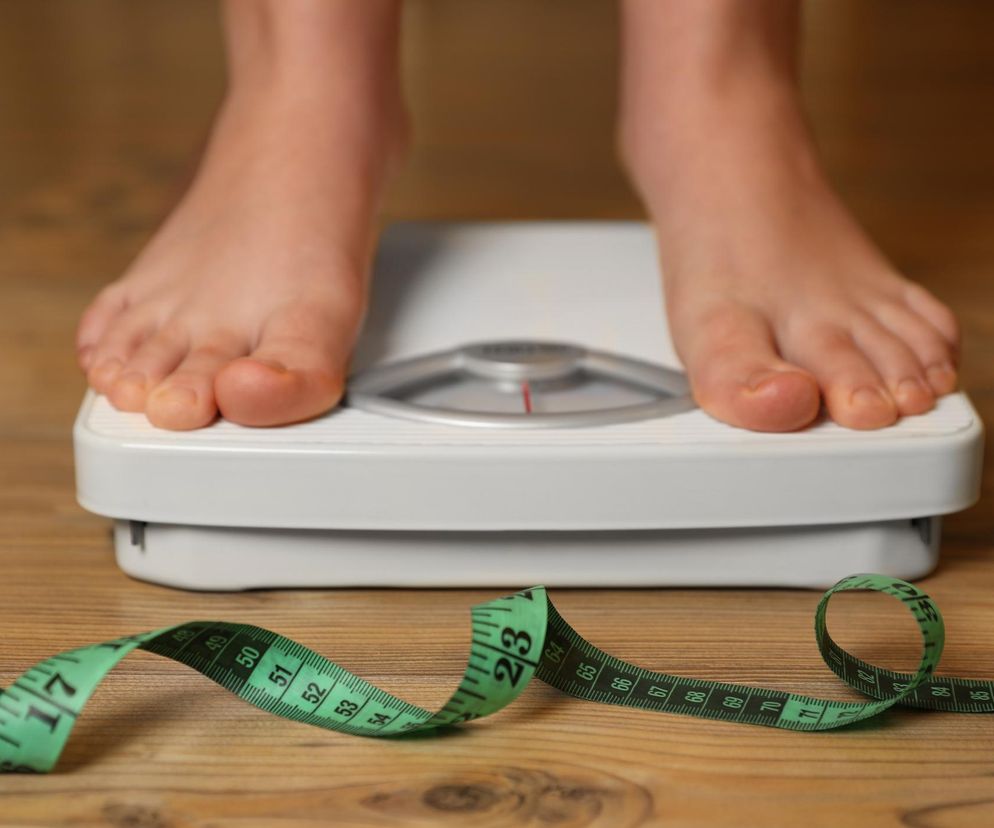 Stopy osoby stojącej na wadze łazienkowej, obok której na drewnianej podłodze leży zwinięta zielona miarka krawiecka. Obraz symbolizuje kontrolę wagi i dbanie o zdrowie, o czym można przeczytać na portalu Poradnik Zdrowie.