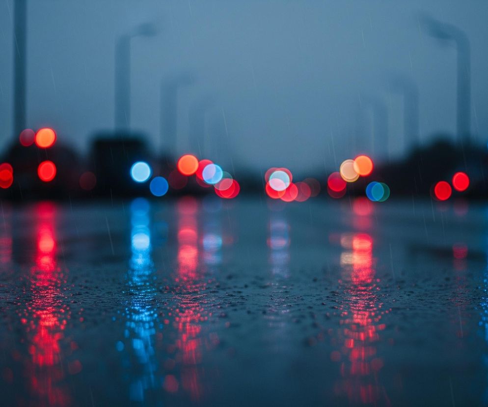 Mokry asfalt w pierwszym planie pokryty jest drobnymi kroplami deszczu, odbijającymi jaskrawe, rozmyte światła w kolorach czerwonym, niebieskim i pomarańczowym. Na środku widoczna jest mokra jezdnia, a w oddali, w rozmazanym tle, widać rzędy drzew i latarni ulicznych, zanurzonych w szarym, mglistym otoczeniu. Padający deszcz tworzy delikatne smugi na całym obrazie, nadając mu zamglony, chłodny charakter, z dominującymi odcieniami błękitu, szarości i granatu.